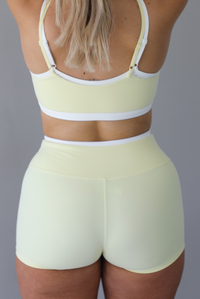 Woman wearing a yellow and white two piece athleisure set featuring a sports bra and biker shorts that have a layered look with the white peeking out from under the yellow. The photo is taken indoors against a white wall and shows a closeup of the back of the shorts. 