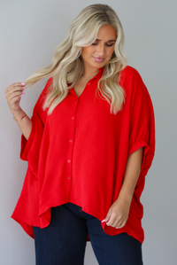 A woman wearing a bright red oversized short-sleeve blouse with functional button closures in the front and dark wide-leg jeans, standing indoors against a white wall.