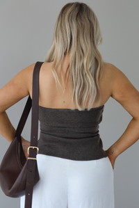 Woman wearing a strapless brown top with non-functional button closures in the front with  a split hem detail, and high waisted, flared hem white linen pants with a brown shoulder bag. The photo is taken indoors against a white wall and shows a closeup of the back of the brown top. 