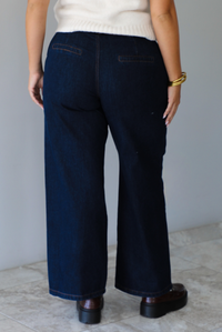 A woman shown from behind wearing dark denim wide-leg jeans, a cream knit top, and brown leather shoes, standing indoors against a white wall with white marble flooring. 
