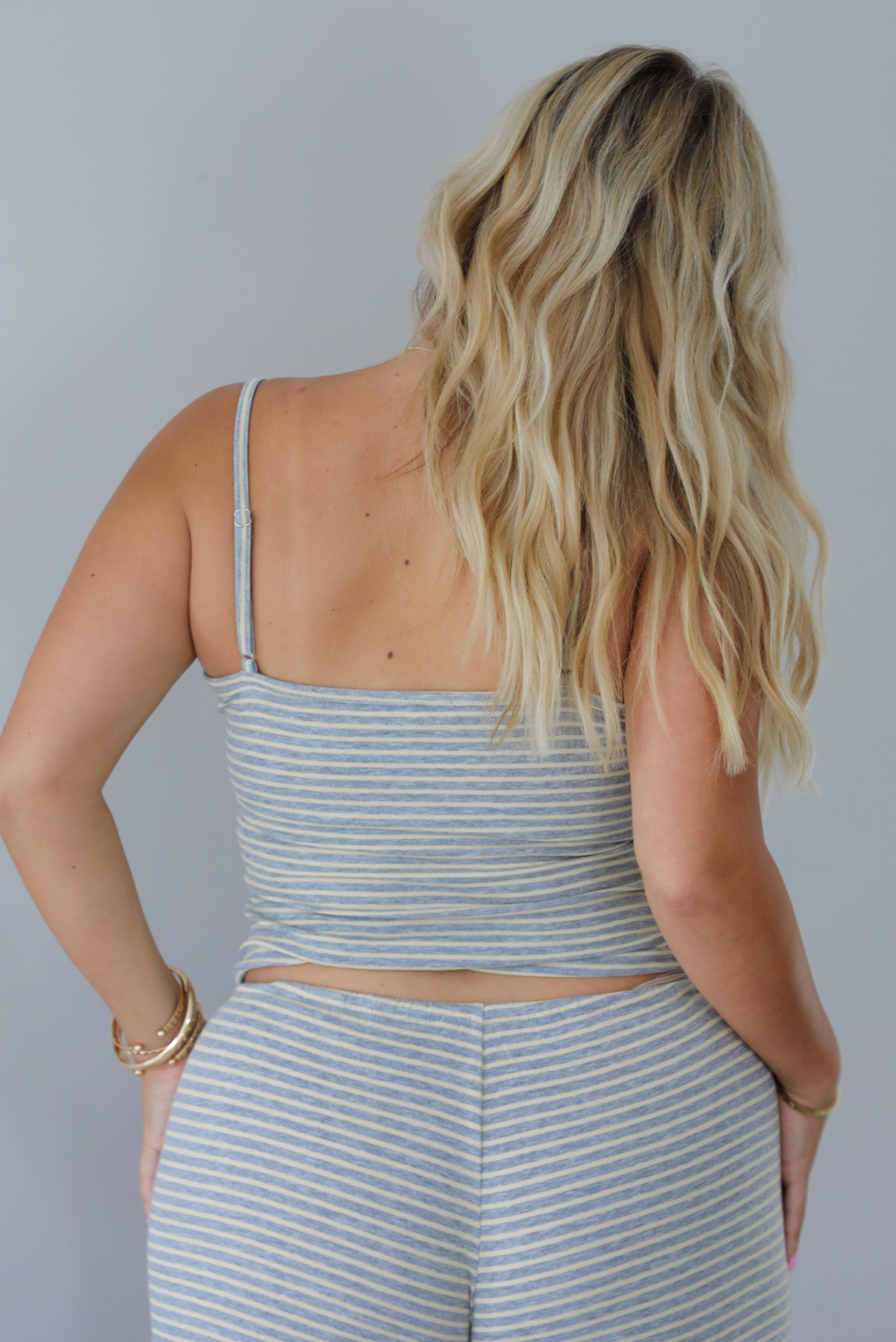 A blonde woman shown from behind modeling a matching two-piece loungewear set consisting of a gray and pale yellow striped crop tank top and coordinating drawstring shorts, set indoors against a white wall. The tank top has adjustable straps. 