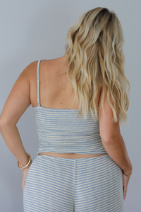 A blonde woman shown from behind modeling a matching two-piece loungewear set consisting of a gray and pale yellow striped crop tank top and coordinating drawstring shorts, set indoors against a white wall. The tank top has adjustable straps. 