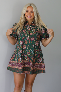 A woman wearing a dark green dress that has a mauve and multicolored floral print pattern throughout, has puffed sleeves, a collared neckline with functional gold button closures in the front, standing indoors against a white wall.