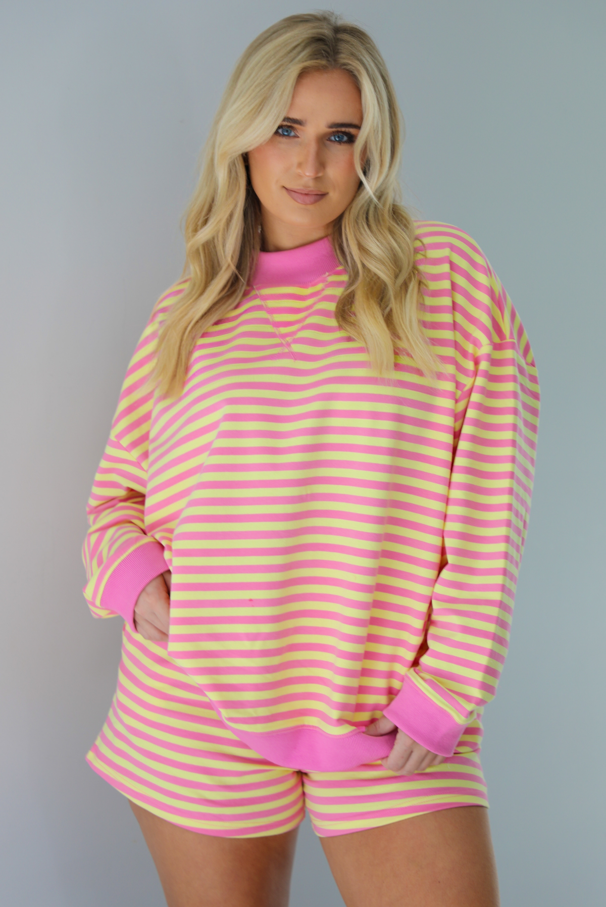 Woman wearing a pink and yellow striped set featuring an oversized sweatshirt and shorts. The photo is taken indoors against a white wall and shows a closeup of the top of the set, which is untucked to show the full length. 