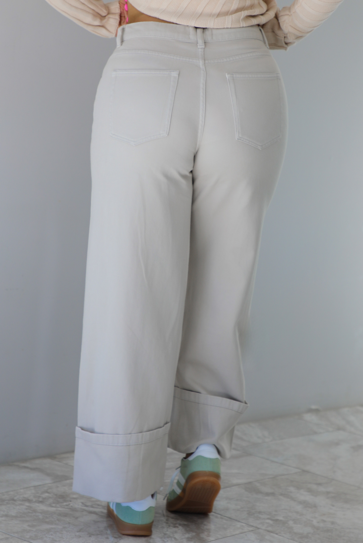 Woman wearing high waisted, sand colored denim pants that have a straight leg cut with a cuffed hem. The photo shows the back of the pants. The photo is taken indoors against a white wall with white marble flooring. 