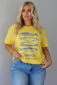 A woman wearing a bright yellow graphic t-shirt with blue sardine illustrations and the text 'Sardines LISBON FISH MARKET', paired with blue jeans, standing indoors against a white wall.