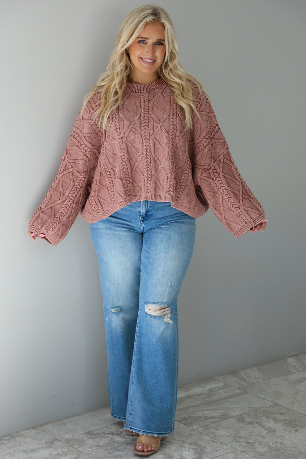 A woman wearing a mulberry oversized sweater paired with a pair of medium wash distressed denim, standing indoors against a white wall with white marble flooring. 