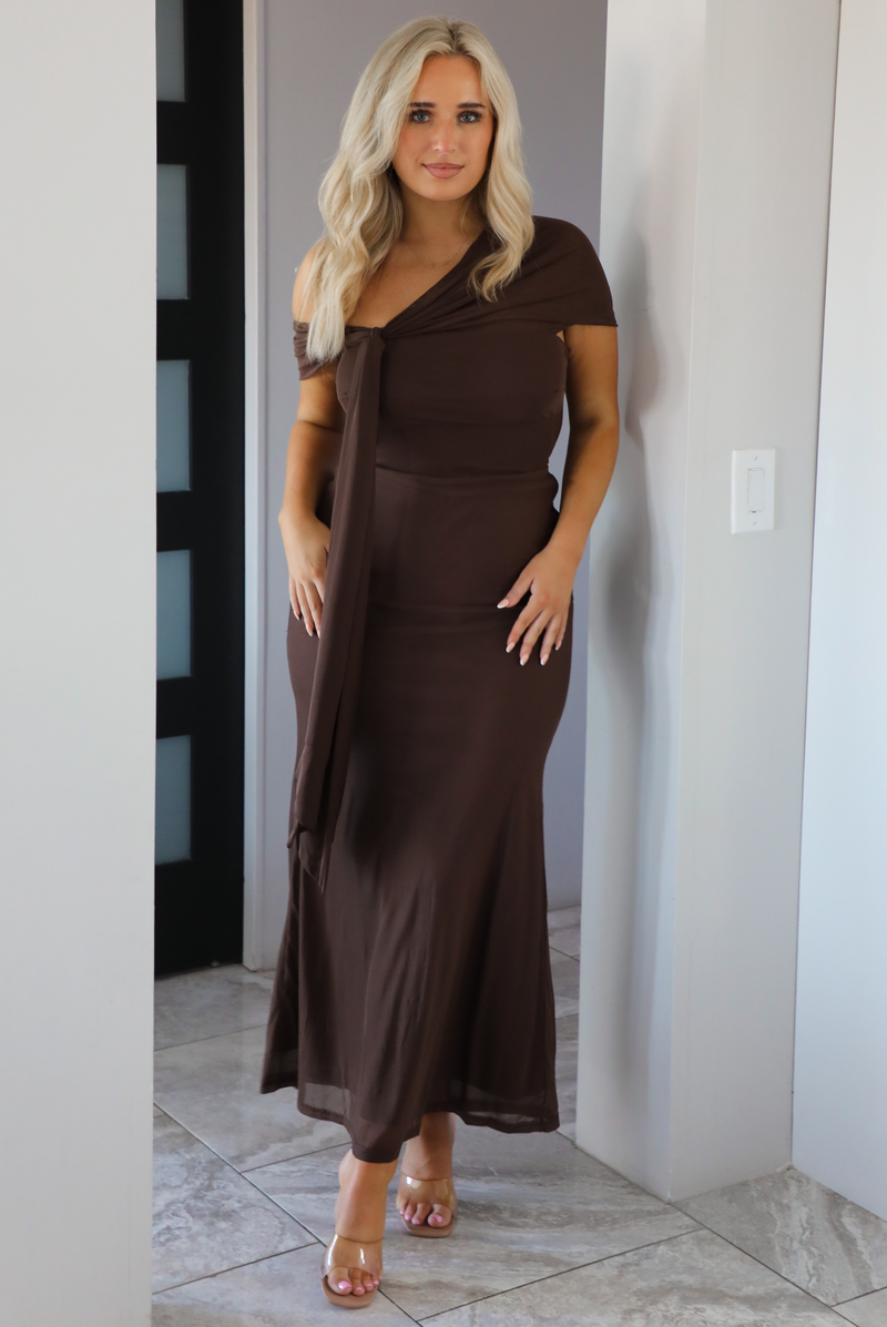 Woman model wearing a chocolate brown formal style dress that's fitted at the bodice, has asymmetrical sleeves, and a mermaid style skirt that's fitted at the hips and flared from the bottom of the high to the hem. The photo is taken inside a modern home in an archway.