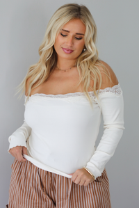 A closeup of a woman wearing a cream off the shoulder long sleeved top, untucked to show the full length, into a pair of camel and cream striped pants, standing indoors against a white wall. 