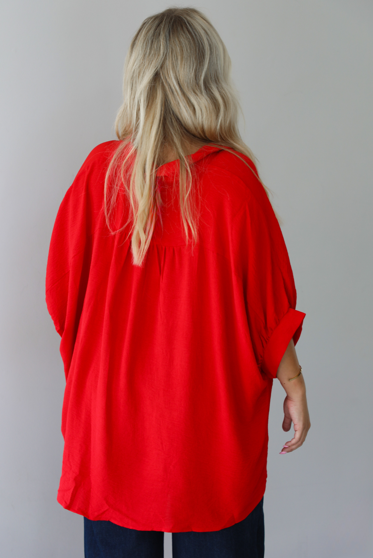 A woman shown from the back wearing a bright red oversized short-sleeve blouse with functional button closures in the front and dark wide-leg jeans, standing indoors against a white wall.
