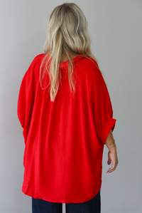 A woman shown from the back wearing a bright red oversized short-sleeve blouse with functional button closures in the front and dark wide-leg jeans, standing indoors against a white wall.
