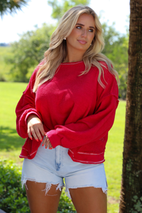 Woman wearing an oversized long sleeved sweater with light wash denim shorts outdoors with greenery in the background. 