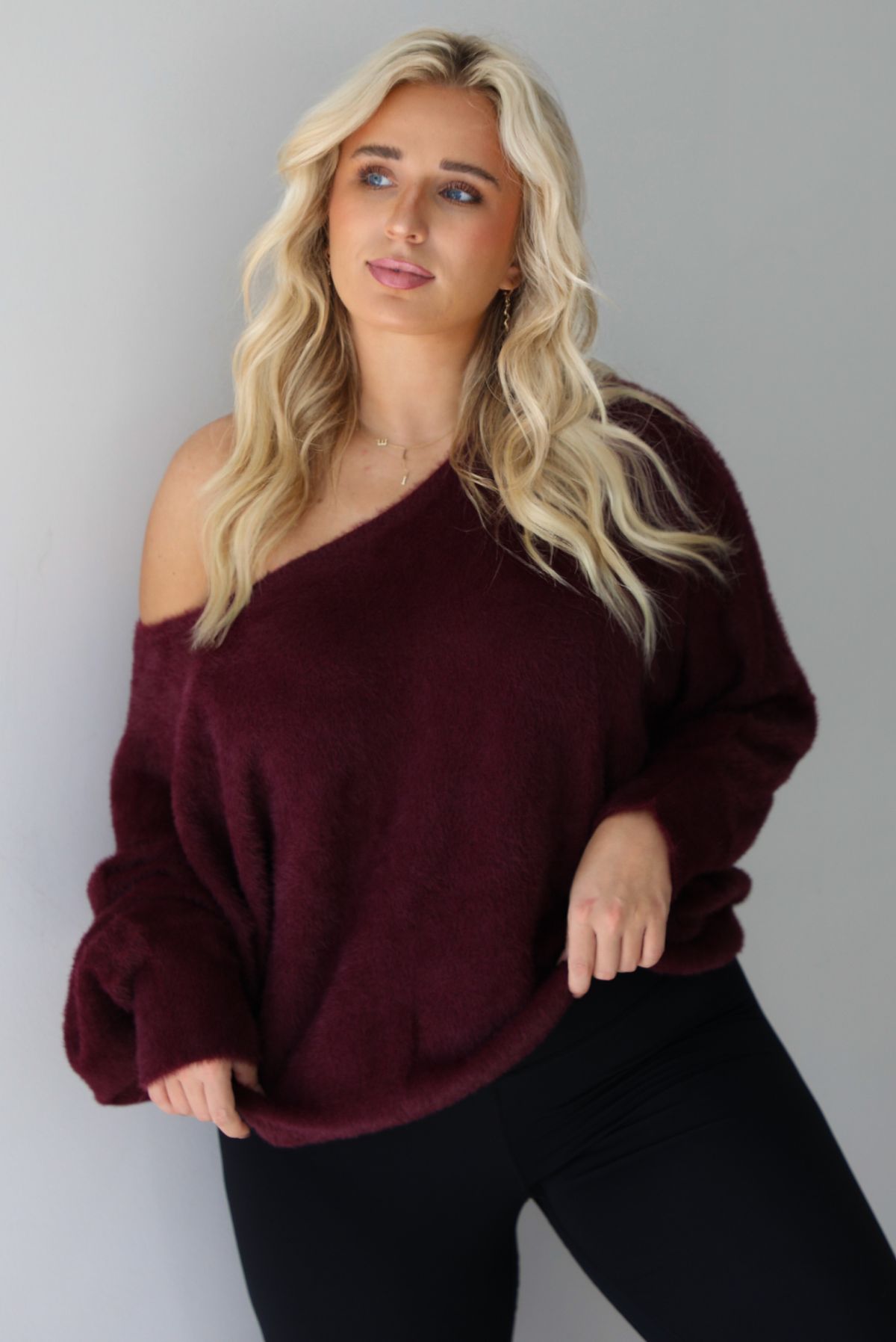 A woman wearing a maroon oversized long sleeved sweater, worn off the shoulder, paired with black leggings, indoors against a white wall.