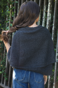 Close up of the back of a woman model outdoors wearing a charcoal grey colored cape cardigan with functional button closures in the front and dark wash denim. 