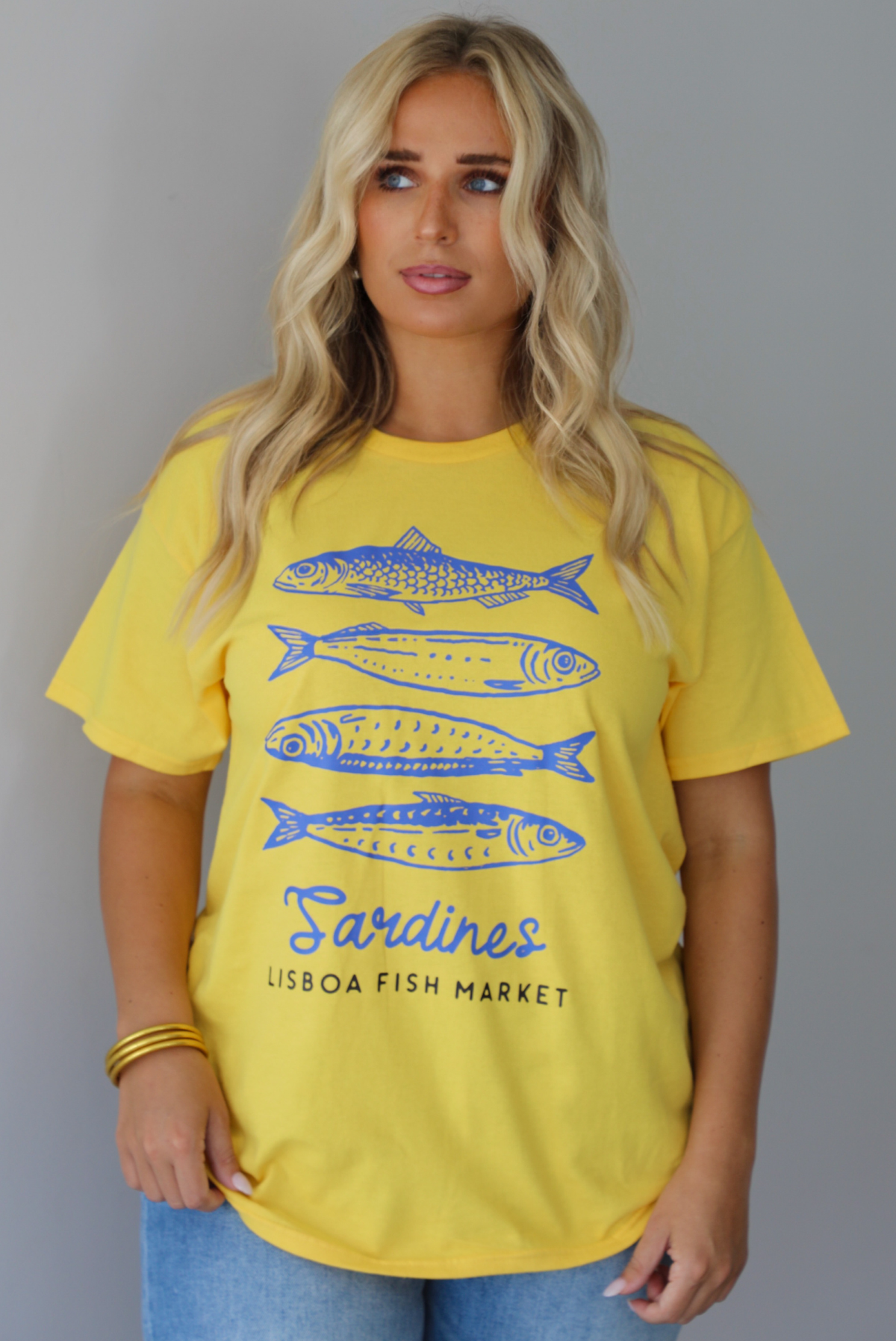 A woman wearing a bright yellow graphic t-shirt with blue sardine illustrations and the text 'Sardines LISBON FISH MARKET', paired with blue jeans, standing indoors against a white wall.
