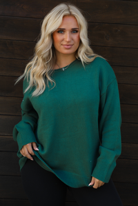 Woman model wearing a green oversized sweater with black leggings, white socks, and tan Uggs. The photo is taken outdoors against a dark brown door. The photo is taken up close and showcases the sweater. 
