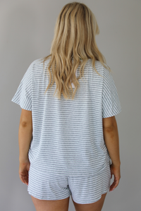 Woman wearing a heather grey and white striped two piece oversized tee and shorts set with green trim at the top of the neckline and on the waistline. The photo is taken indoors against a white wall and shows the back of the top. 