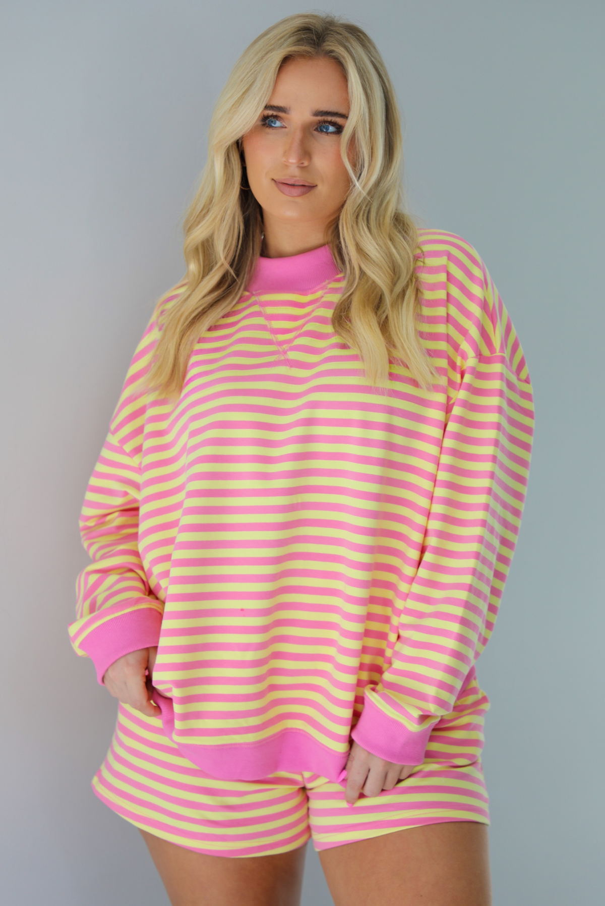 Woman wearing a pink and yellow striped set featuring an oversized sweatshirt and shorts. The photo is taken indoors against a white wall and shows a closeup of the top of the set, which is untucked to show the full length. 