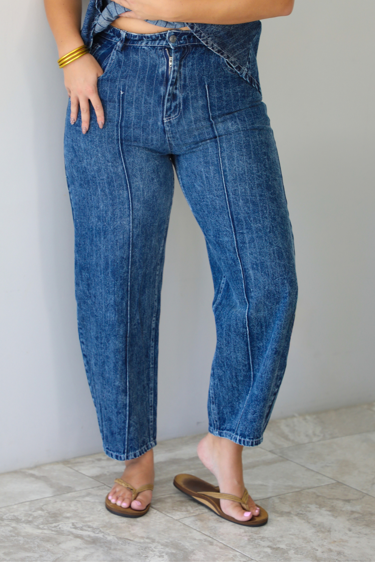 A woman wearing a matching two piece dark wash denim set with a pin striped design on it featuring a sleeveless peplum style top and high waisted denim jeans, standing indoors against a white wall with white marble flooring. The photo focuses on the pants only, which have functional pockets on either side with a button and zipper closure at the top of the pants. 