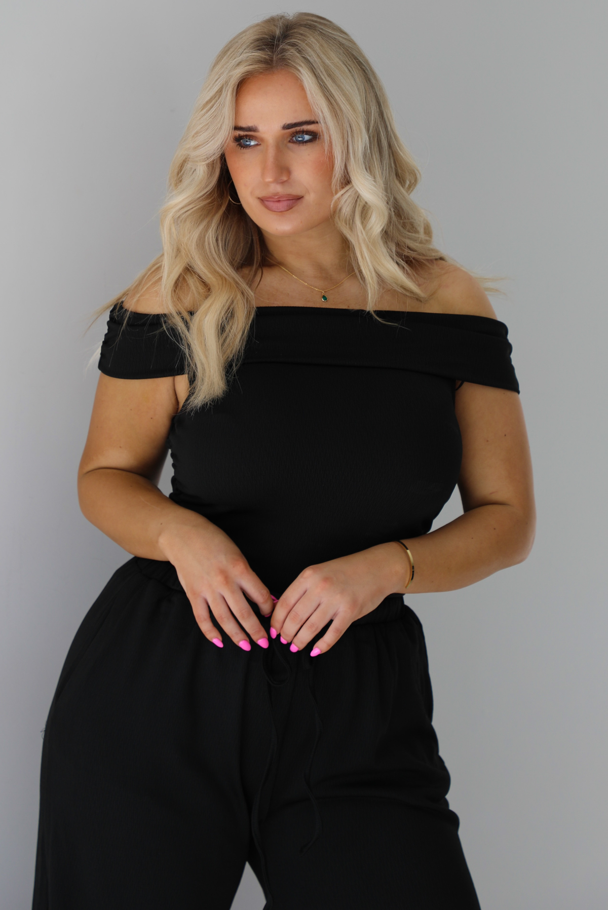 Woman wearing a two piece set featuring an off the shoulder top and high waisted pants. The photo shows a closeup of the top. The photo is taken indoors against a white wall.