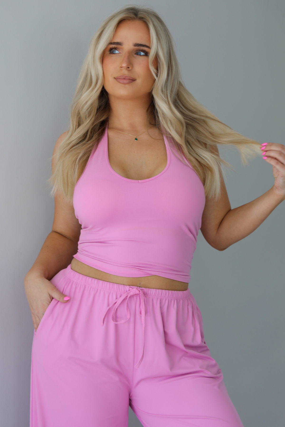 Woman wearing a two pieced pink set featuring a halter top and comfy pants with a flared hem detail. The photo is taken indoors against a white wall, and shows a closeup of the halter top, which. has a slightly cropped cut. 