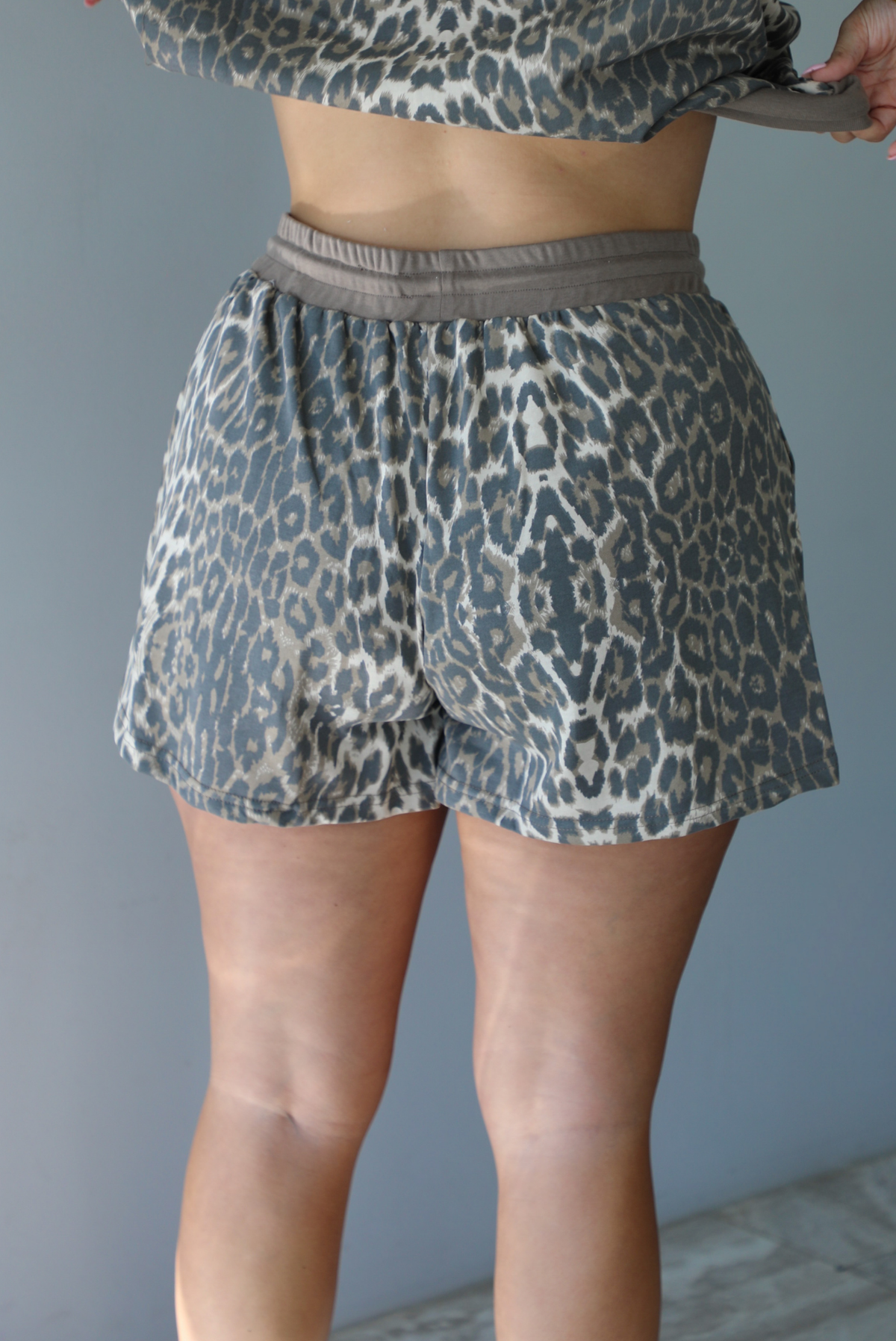 A woman shown from behind modeling a matching leopard-print quarter-zip sweater and shorts set with gray drawstrings, showing a closeup of the back of the shorts, standing indoors against a white wall.