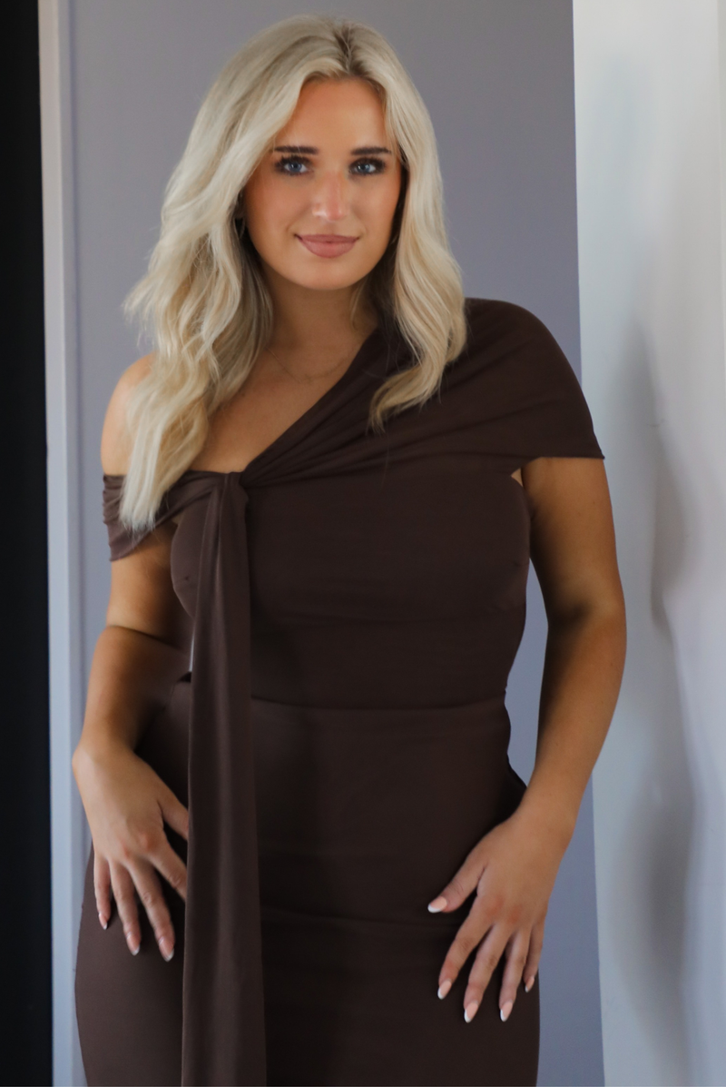 Woman model wearing a chocolate brown formal style dress that's fitted at the bodice, has asymmetrical sleeves, and a mermaid style skirt that's fitted at the hips and flared from the bottom of the high to the hem. The photo is taken inside a modern home in an archway. The photo showcases a close up of the bodice of the dress.