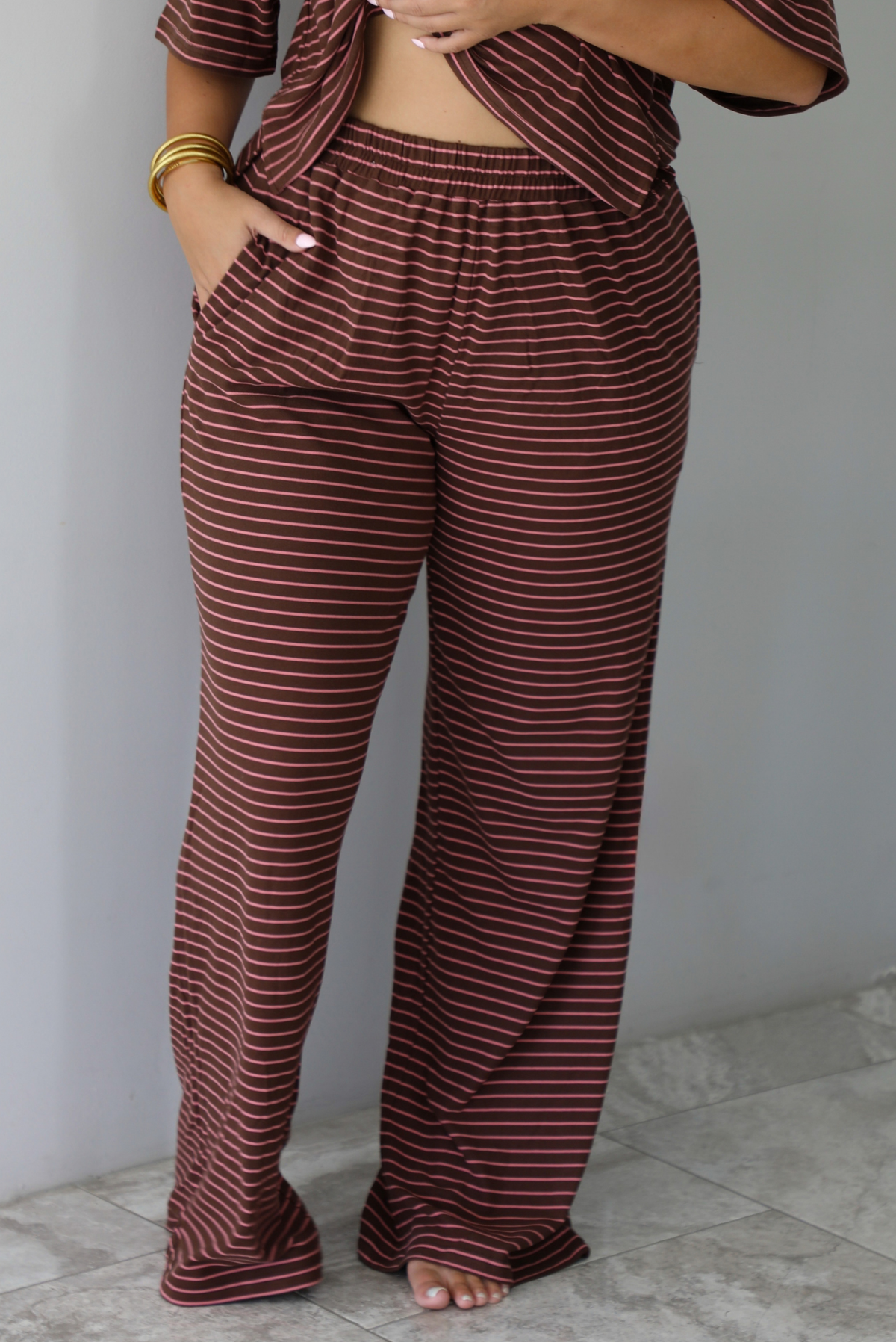 A woman wearing a brown and pink striped two piece set featuring a slightly cropped tee and high waisted pants, standing indoors against a white wall with white marble flooring. The photo shows a closeup of the pants, which have an elastic waistline and functional pockets on either side. 