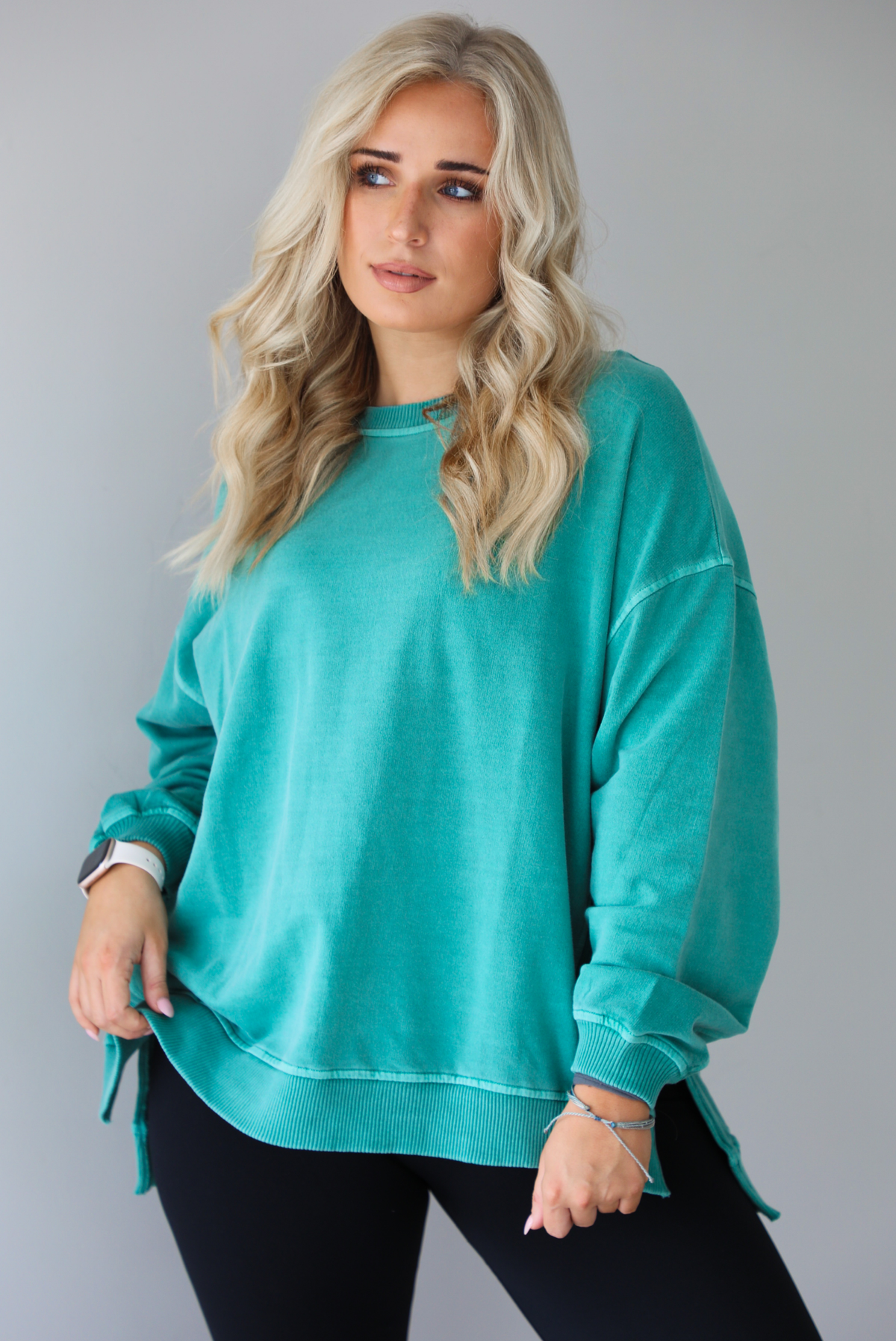 Woman wearing a teal oversized sweater with black leggings that have a slightly flared hem with split hem details in the front. The photo shows a closeup of the sweater and has a white background. 