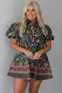 A woman wearing a dark green dress that has a mauve and multicolored floral print pattern throughout, has puffed sleeves, a collared neckline with functional gold button closures in the front, standing indoors against a white wall.