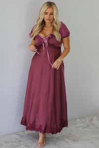 A woman modeling a long, burgundy maxi dress with short puff sleeves, a lace-up sweetheart neckline, and a flowing satin-like skirt, standing indoors against a white wall with white marble flooring. 