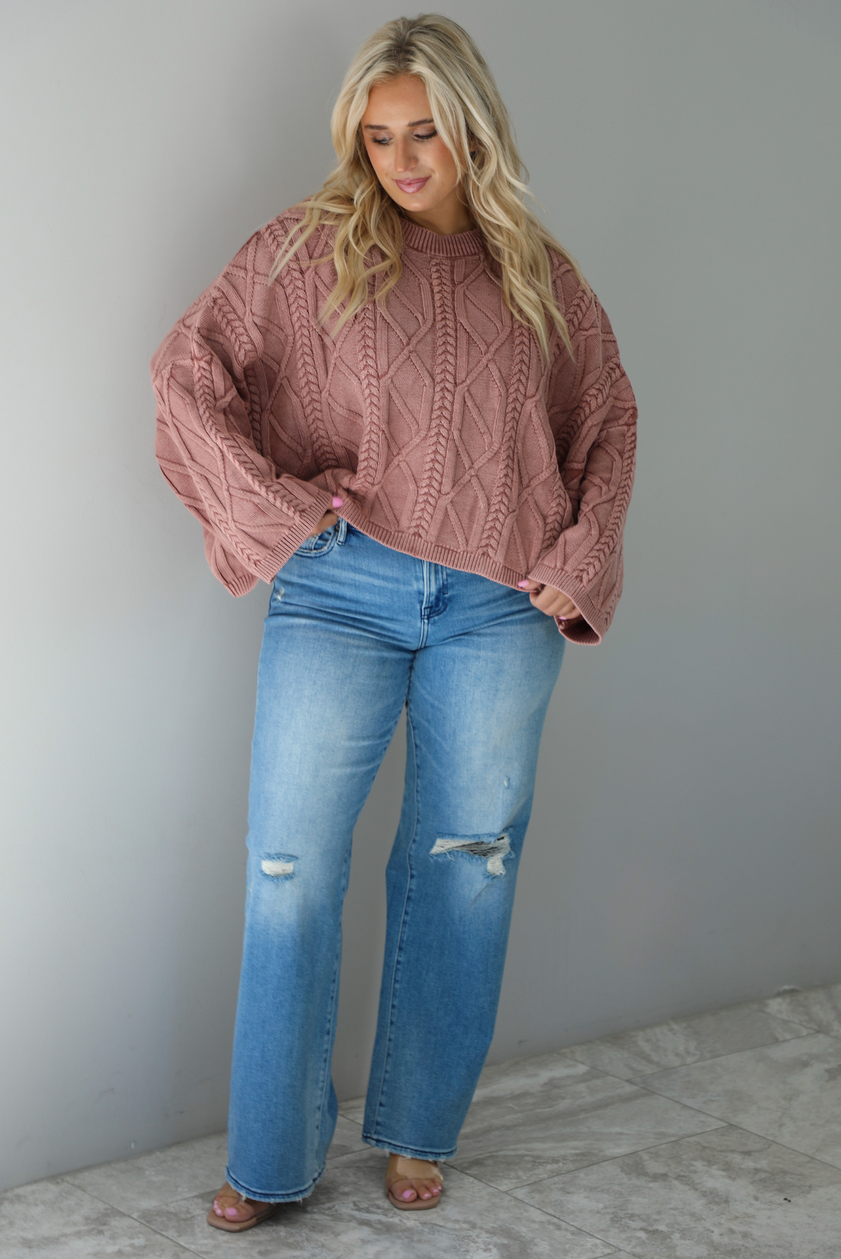 A woman wearing a mulberry oversized sweater paired with a pair of medium wash distressed denim, standing indoors against a white wall with white marble flooring. 