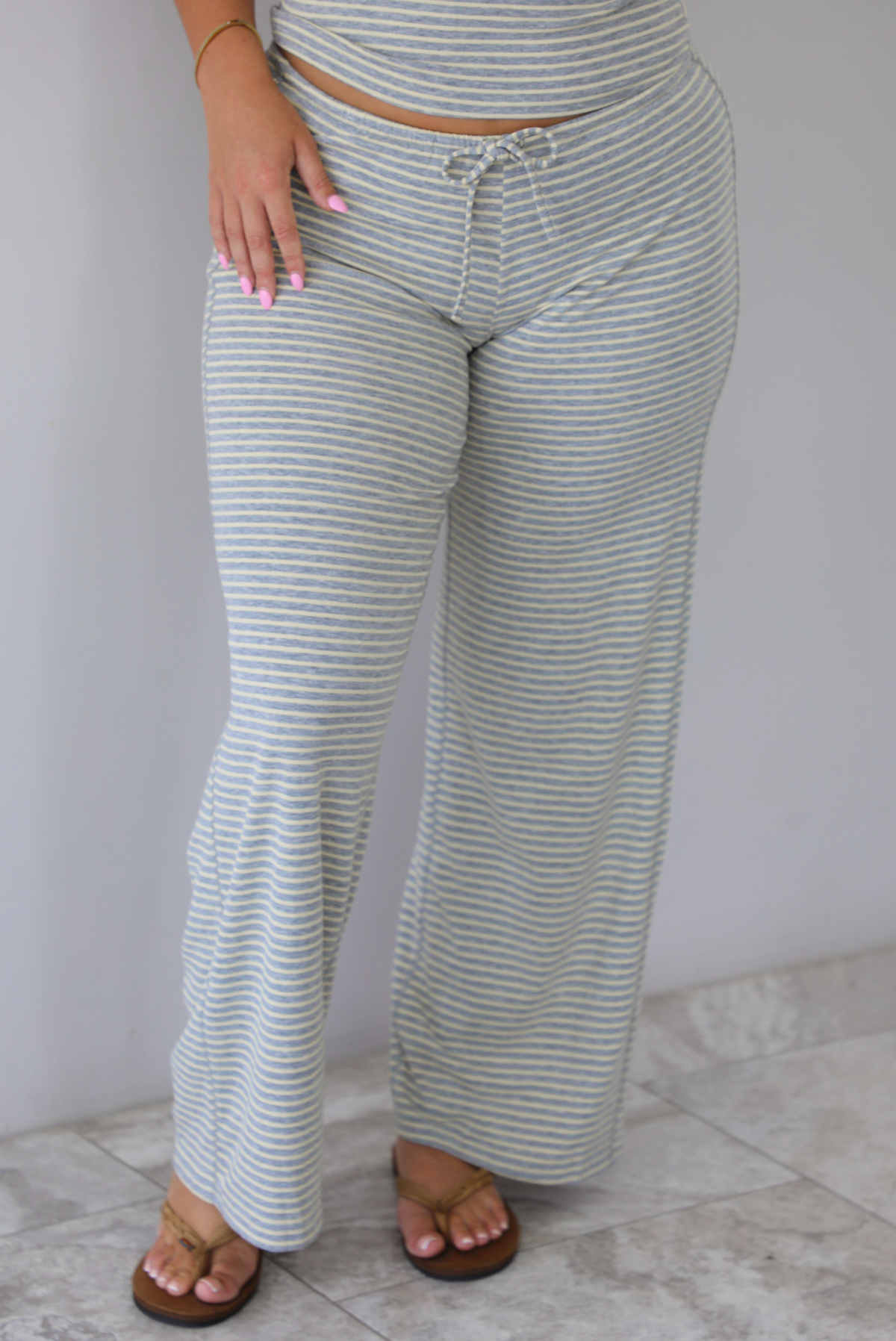 A person wearing light gray and pale yellow horizontally striped wide-leg pants with a drawstring waistband, paired with brown sandals, standing indoors against a white wall with white marble flooring.