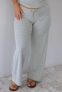 A person wearing light gray and pale yellow horizontally striped wide-leg pants with a drawstring waistband, paired with brown sandals, standing indoors against a white wall with white marble flooring.