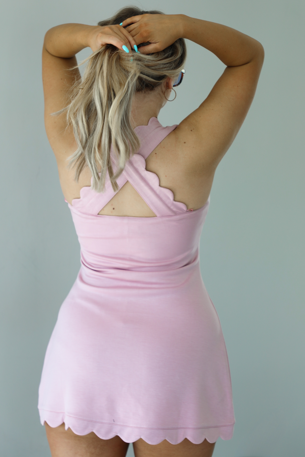 Woman wearing a pink dress with scalloped hem, white sneakers, and sunglasses. The photo is taken in front of a white wall and shows a closeup of the back of the dress, which has a crisscross design with the straps.