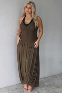 A woman wearing an olive maxi dress with black trim detailing at the v-halter neckline, has a fitted bodice and drop waist design, with a maxi-length flowy skirt, standing indoors against a white wall with white marble flooring. 