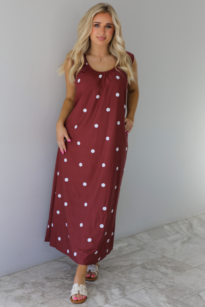 Woman wearing a red brown maxi length dress that has a white polka dot print pattern throughout. The dress is paired with sand sandals. The photo is taken indoors against a white wall with white marble flooring. 