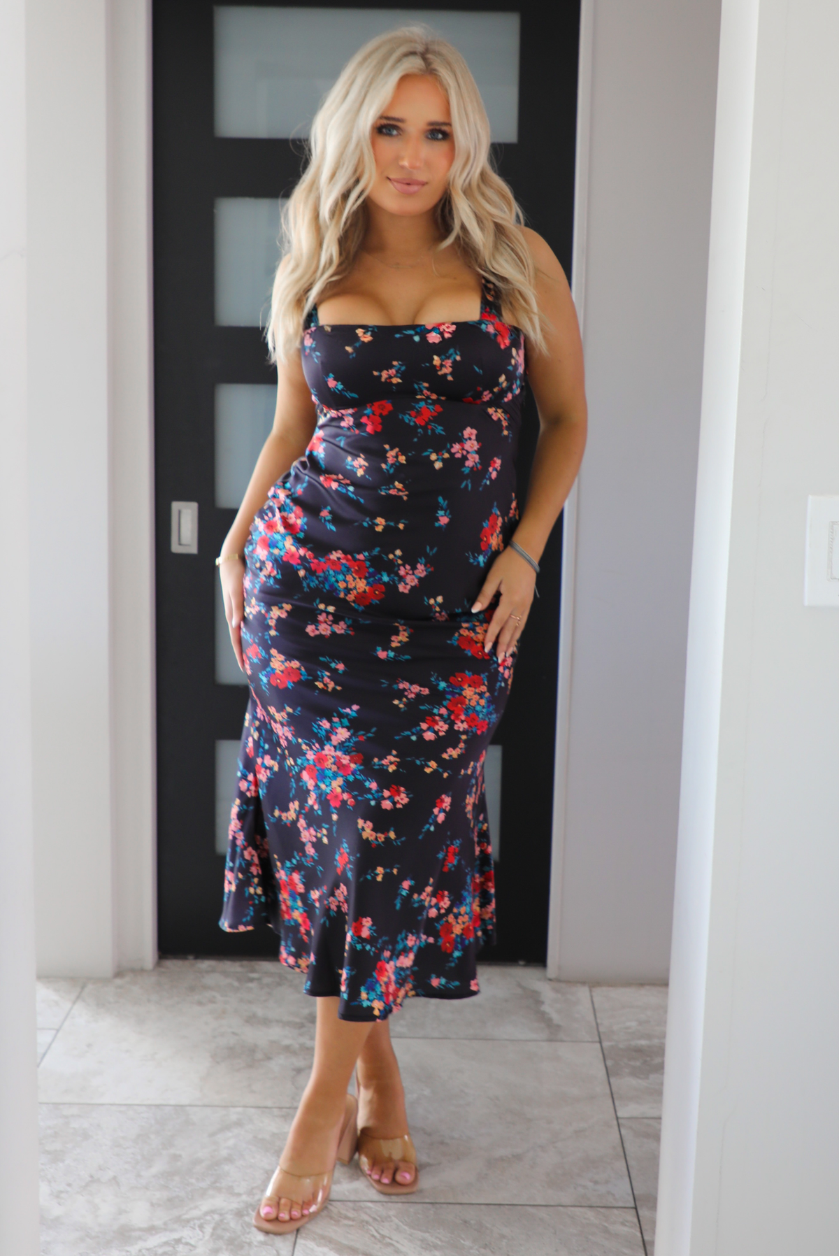 Woman model wearing a navy dress with a multi-colored floral print. The dress is a satin material, is a midi length, and is curve-hugging. The photo is taken inside a modern white and black interior. 