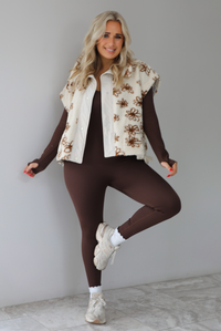 The photo features a model wearing a beige sherpa vest with a brown floral print pattern throughout, functional button closures in the front, and that is oversized with a brown, fitted jumpsuit that's paired with white scrunchie socks and beige sneakers. The photo is taken indoors against a white wall with white marbled flooring. 