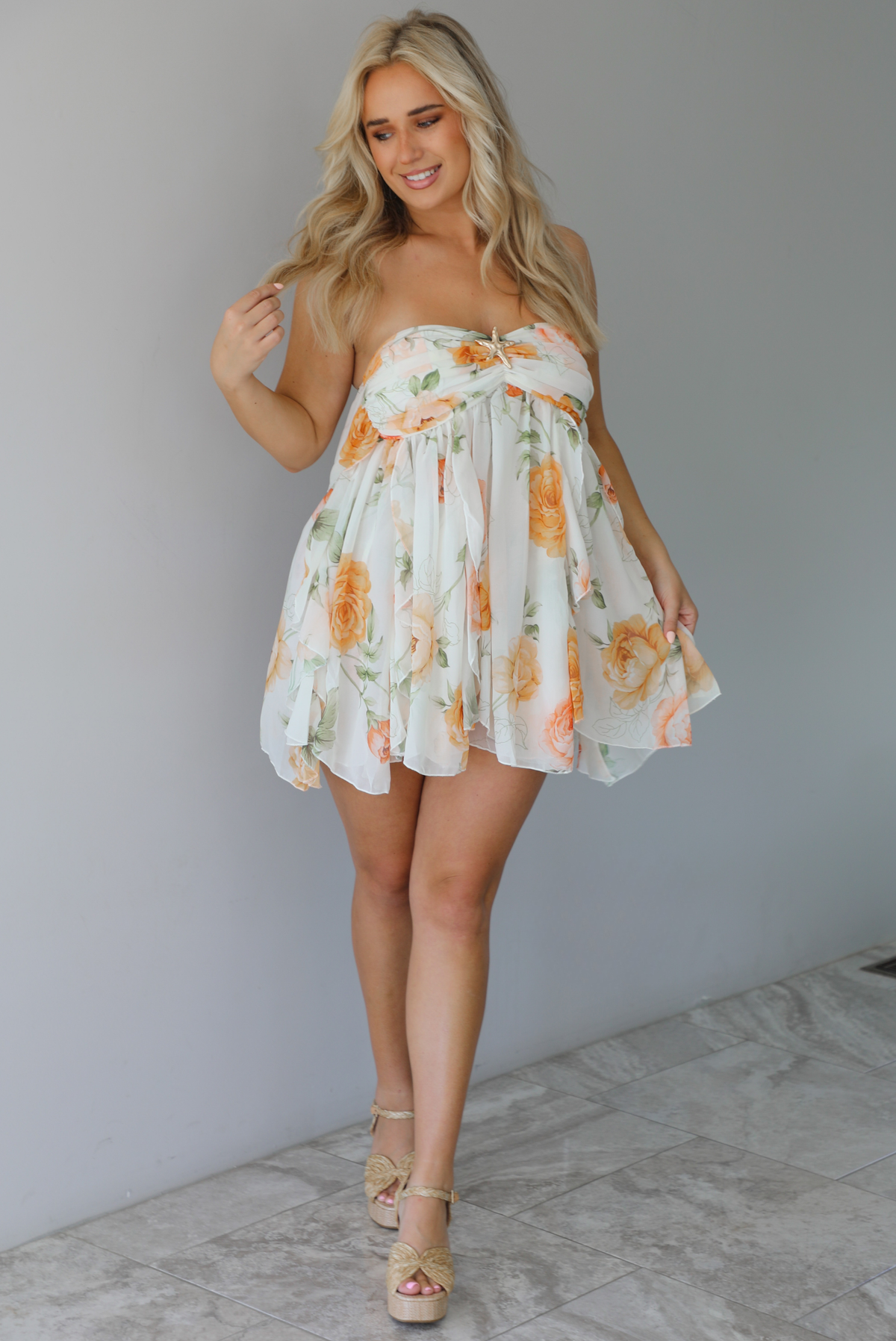 Woman wearing a white, strapless, babydoll cut dress that has an orange and green floral print pattern and flowy skirt indoors against a white wall with white marble flooring. 