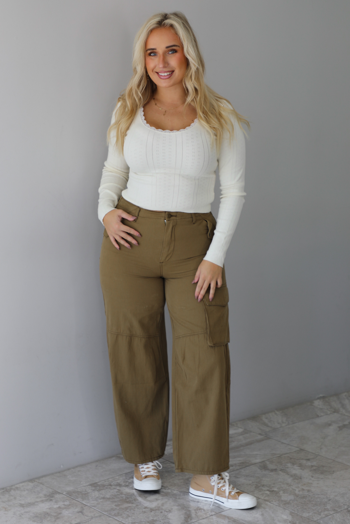 A woman wearing a cream knit long sleeved top and high waisted olive-khaki cargo pants, standing indoors against a white wall with white marble flooring. 