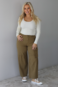 A woman wearing a cream knit long sleeved top and high waisted olive-khaki cargo pants, standing indoors against a white wall with white marble flooring. 