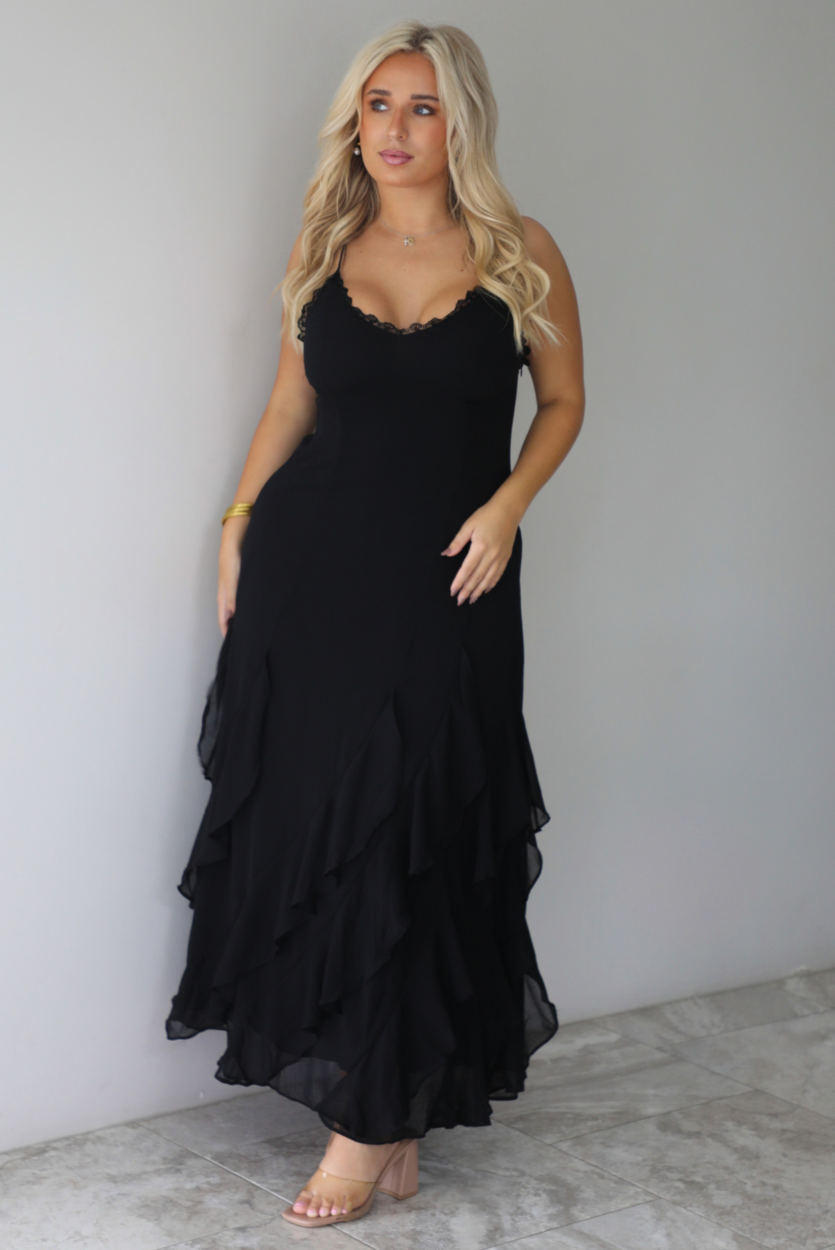 A woman wearing a black formal maxi dress with lace trim detailing at the bodice and ruffled details at the skirt, standing indoors against a white wall with white marble flooring. 