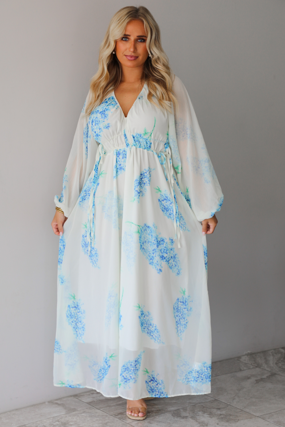 A woman modeling a white maxi dress with a blue floral print and a sheer, flowing overlay, standing indoors against a white wall with white marble flooring. 