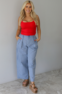 Woman wearing a red tank top high waisted navy and white plaid pants indoors against a white wall with white marble flooring. 