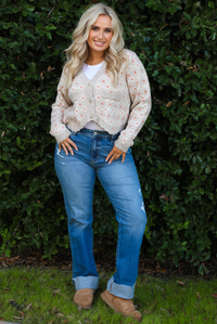 Woman model wearing a grey cardigan that has an orange and white flora print pattern throughout, long sleeves, an asymmetrical hem, and functional button closures in the front, with a long sleeved white top, medium wash denim jeans, and brown shoes in front of a green hedge outdoors. 