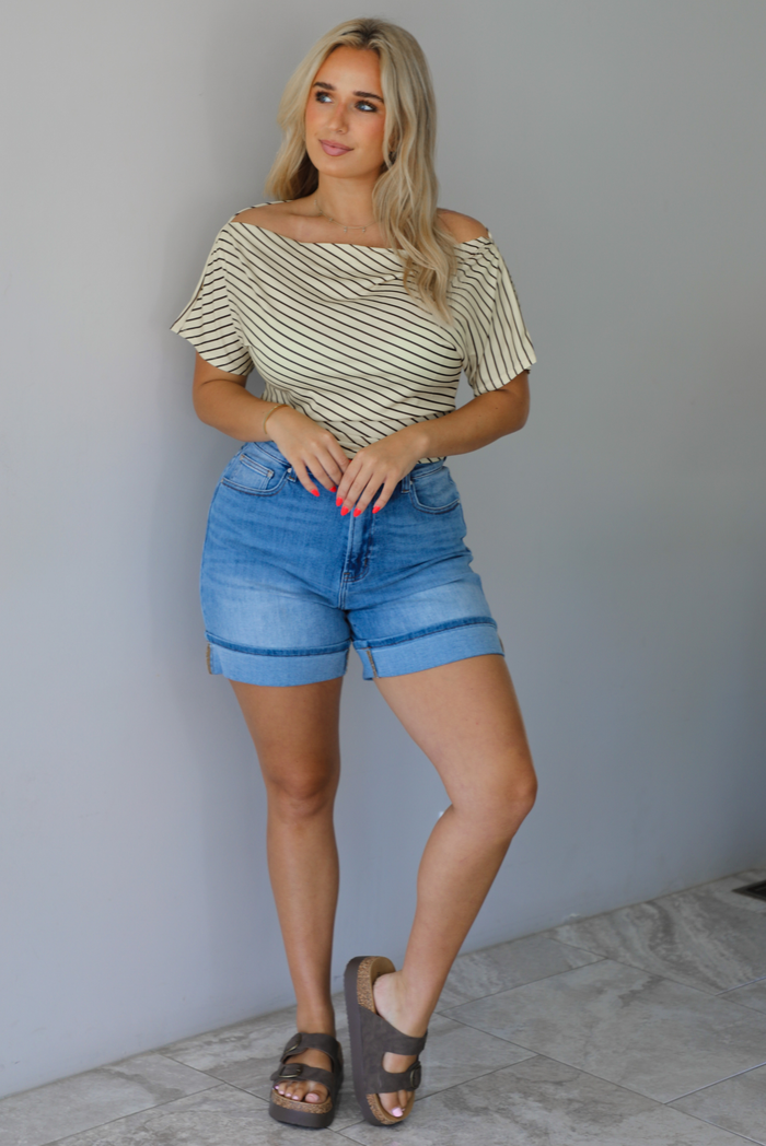 Woman wearing a pale yellow top with a taupe striped pattern with medium wash denim shorts indoors against a white wall with white marble flooring.