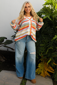 Woman model wearing an orange, lavender, yellow, muted green, blue, and white striped oversized cardigan with functional button closures in the front and an oversized fit with a light wash flared hem denim. The photo is taken outdoors against a white wall and greenery in the back. 