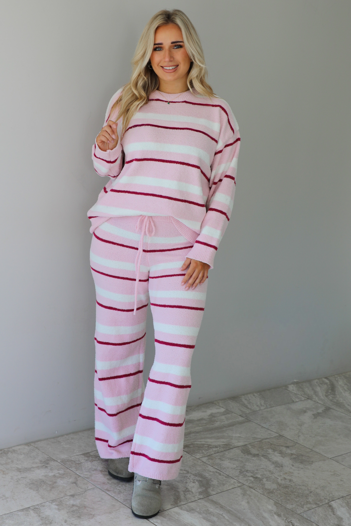 Woman wearing a pink set with a darker pink and white striped design. The set consists of an oversized sweater top and high waisted pants. The sweater of the set is tucked slightly in the front of the pants. The photo is taken indoors against a white wall with white marble flooring. 