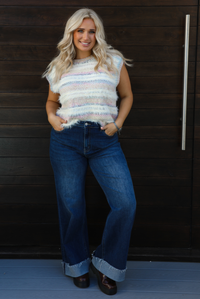 Woman model wearing a soft, striped sweater vest with dark wash denim jeans. The photo is taken outdoors against a dark brown wooden door. 
